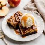 Two apple crumble bars with caramel drizzle and ice cream on a plate.