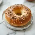 A mouthwatering high protein bagel sitting on a simple white ceramic plate, golden brown and fluffy.