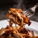 A fork lifting juicy, saucy shredded BBQ chicken from a white plate.