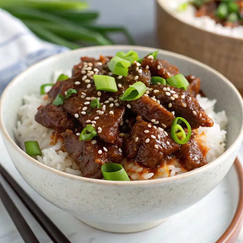 A bowl of sticky Korean beef over rice topped with sesame seeds and green onions.