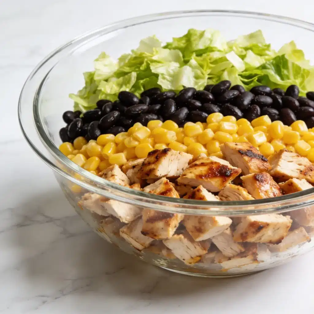 A glass bowl showing the layers and assembly of a high protein bbq chicken chopped salad.