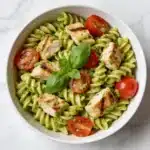 A top-down view of a creamy High Protein Chicken Pesto Pasta Salad with grilled chicken and cherry tomatoes in a white bowl.