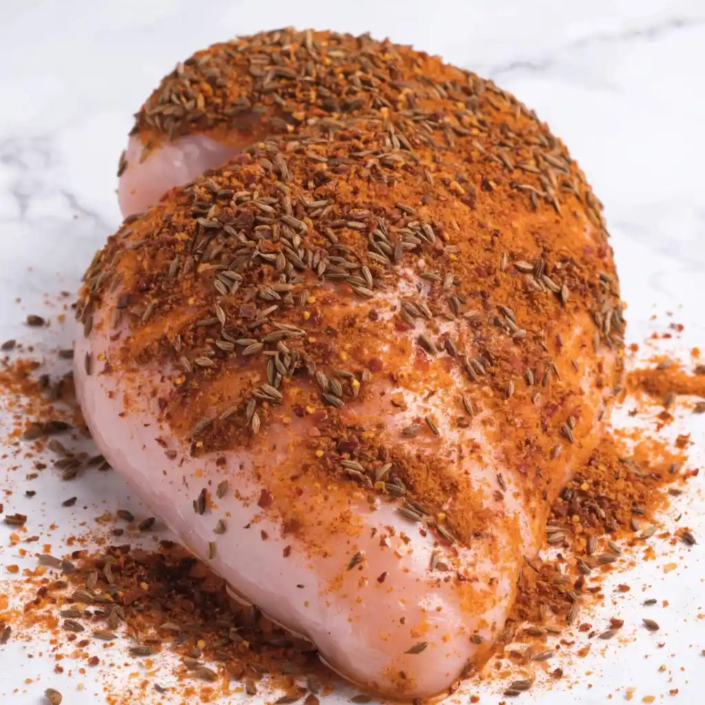 A close-up of raw chicken breast being heavily seasoned with a cumin spice blend.
