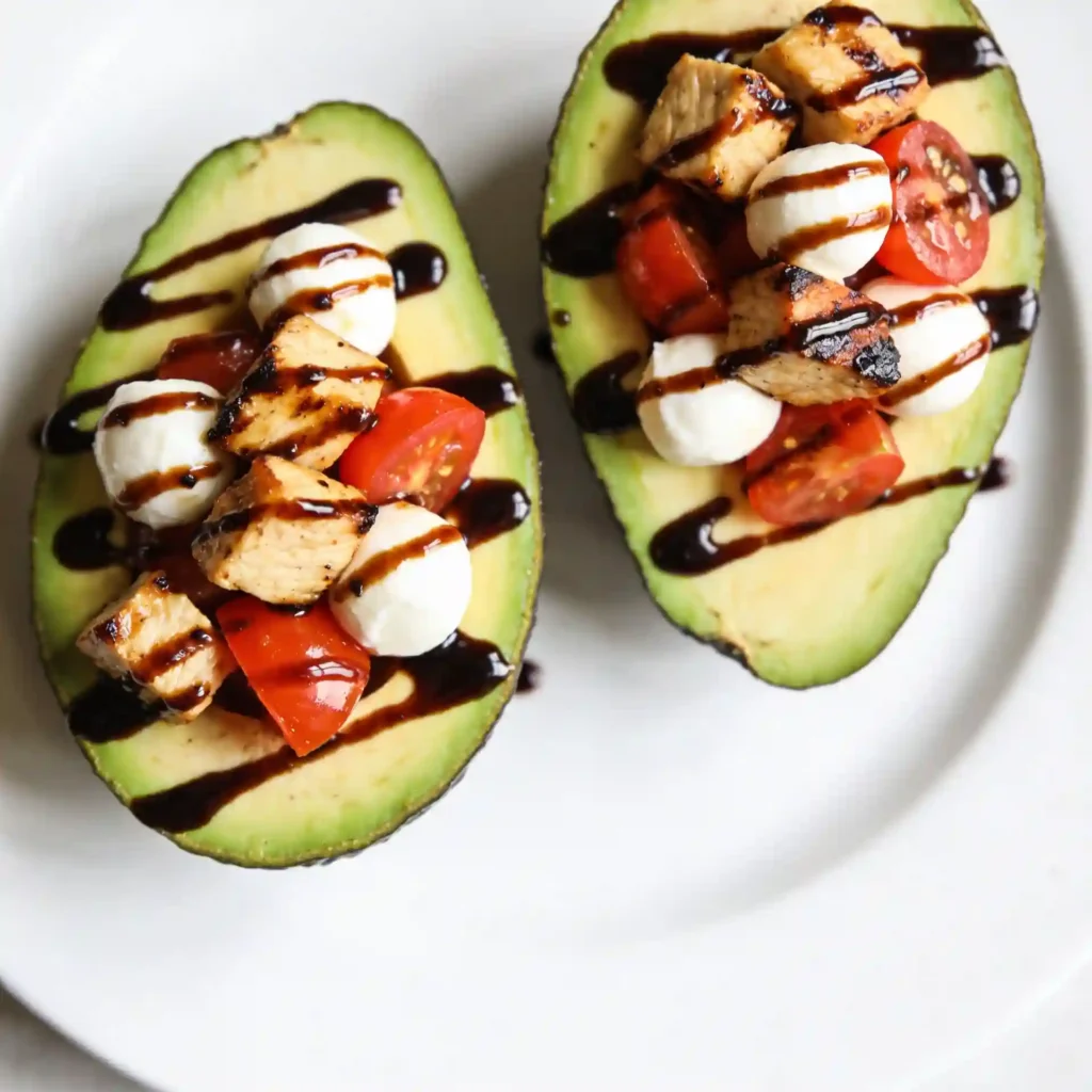 Two avocado halves stuffed with chicken caprese salad and drizzled with balsamic glaze on a white plate.