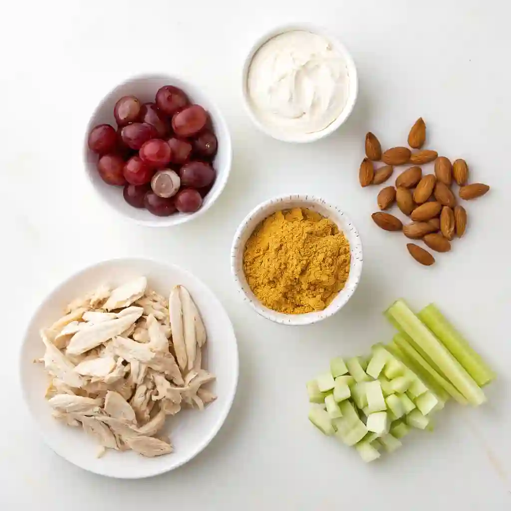 An organized flat-lay of chicken, Greek yogurt, curry powder, grapes, celery, and almonds on a white background.