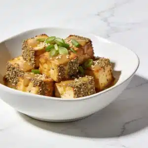 A white bowl featuring golden, crispy Hemp Crusted Tofu drizzled with a glossy miso ginger glaze.