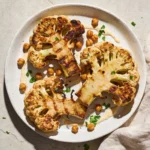 A white plate featuring a deeply caramelized Roasted Cauliflower Steak topped with chickpeas, tahini, and pomegranate seeds.