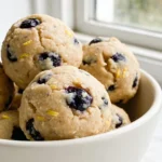 A white bowl of no bake blueberry muffin protein balls speckled with dried blueberries.