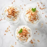 Three elegant glass tumblers filled with creamy white coconut cottage cheese mousse, garnished with toasted coconut flakes on a white quartz counter.