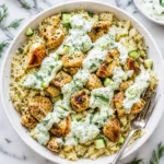 A close-up top-down view of a white bowl filled with chicken, rice, and a heavy drizzle of creamy cucumber dill tzatziki sauce.