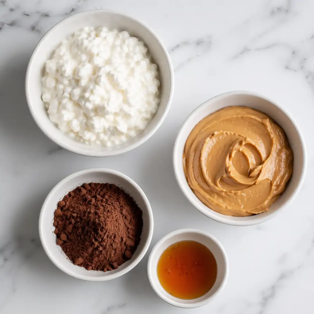 Small modern white bowls filled with cottage cheese, peanut butter, cocoa powder, and maple syrup on a marble counter.