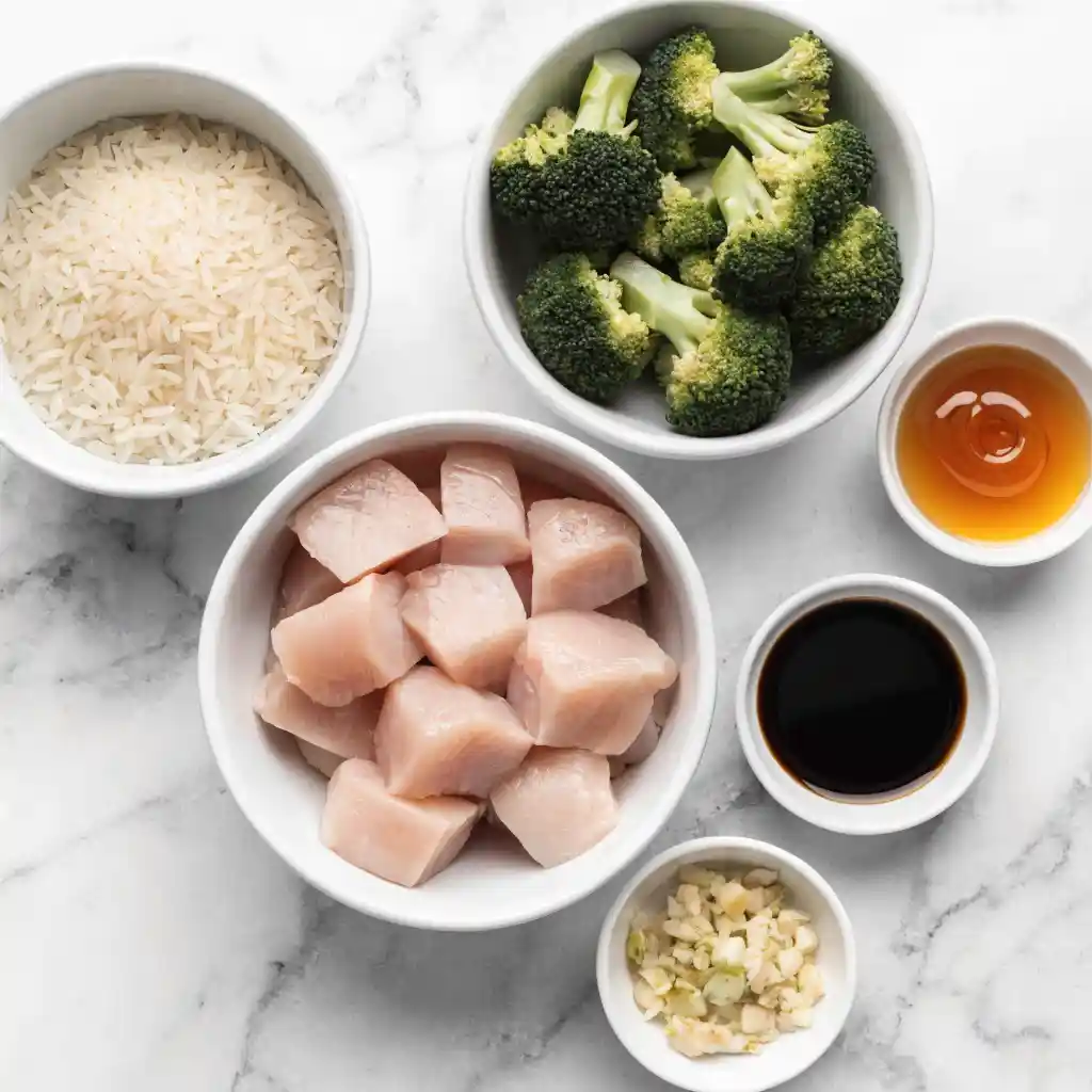 Small white bowls filled with raw chicken, broccoli, rice, honey, soy sauce, and garlic.