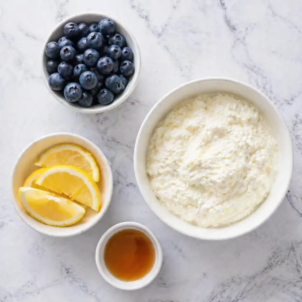 Small modern white bowls filled with cottage cheese, fresh lemons, maple syrup, and blueberries on a marble counter.