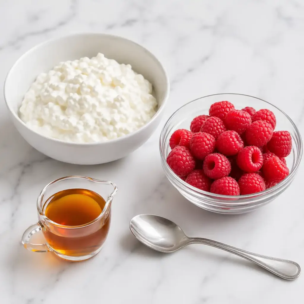 A bowl of thick cottage cheese, fresh red raspberries, and a glass of maple syrup arranged neatly on a white marble surface.