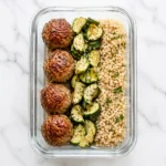 A glass meal prep container filled with high protein turkey meatballs organized in neat rows on a marble counter.
