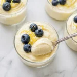 A macro overhead photograph sharply focused on the creamy, velvety texture of lemon cottage cheese mousse in an elegant glass tumbler with a silver spoon and blueberries.