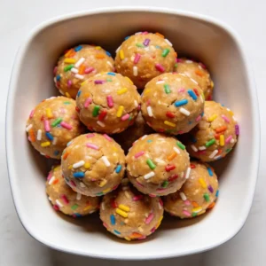 An overhead view of a modern white bowl filled with no bake birthday cake protein balls covered in rainbow sprinkles.