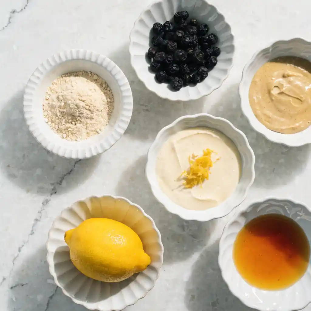 Small white bowls filled with almond flour, dried blueberries, cashew butter, lemon, and maple syrup.