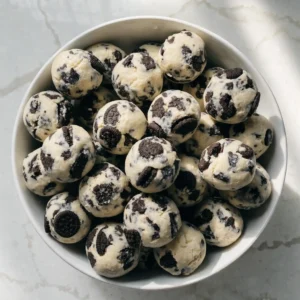 An overhead view of a modern white bowl filled with pale cookies and cream protein balls loaded with dark chocolate cookie chunks.