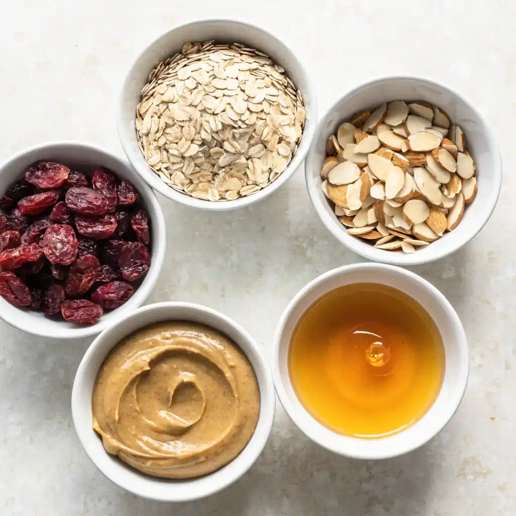 Small white bowls filled with raw oats, dried cranberries, sliced almonds, almond butter, and honey.