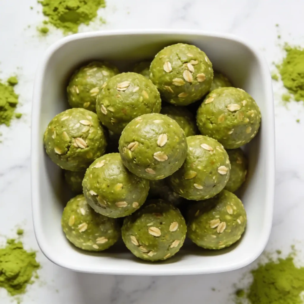 An overhead view of a modern white bowl filled with vibrant Matcha Green Tea Energy Balls, styled with a bamboo matcha whisk.