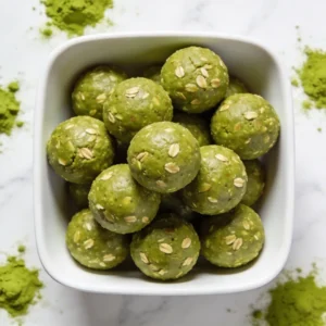 An overhead view of a modern white bowl filled with vibrant Matcha Green Tea Energy Balls, styled with a bamboo matcha whisk.