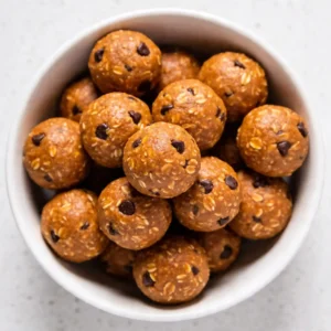 An overhead view of a modern white bowl filled with Pumpkin Spice Protein Balls, styled with a white pumpkin and cinnamon sticks.