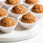 A small cluster of smooth carrot cake bites in white cupcake liners on a glossy white surface with a cinnamon stick.