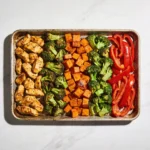 A top-down view of a sheet pan chicken and veggies meal prep with ingredients arranged in colorful columns.