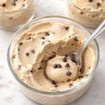 A close-up macro shot of a silver spoon scooping thick, perfectly smooth pale golden cookie dough cottage cheese mousse with mini chocolate chips from a glass tumbler.