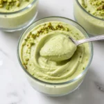 A close-up macro shot of a silver spoon scooping thick, perfectly smooth swirled pale green pistachio cottage cheese mousse from a glass tumbler.